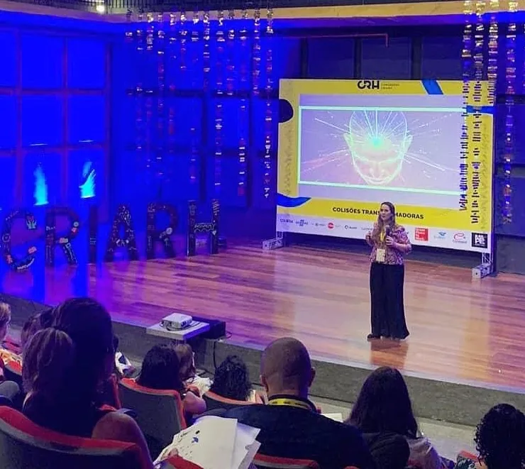 Cristiane Barreto sozinha no palco principal do CRIARH, em Recife, sob luz azul, apresentando a palestra Colisões Transformadoras para uma plateia sentada