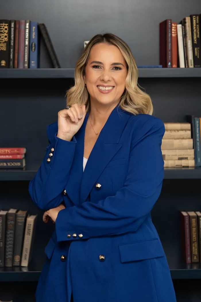 Cristiane Barreto de pie junto a un sillón, con blusa azul claro, y un cuadro geométrico abstracto en la pared detrás de ella