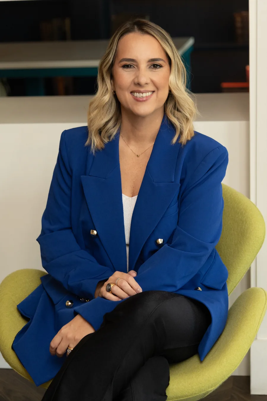 Cristiane Barreto, PhD, sitting in a lime-green armchair in a royal blue blazer at her consulting office