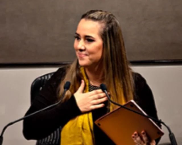 Cristiane Barreto à mesa de oradores no One World Trade Center, com a mão no coração enquanto fala ao público