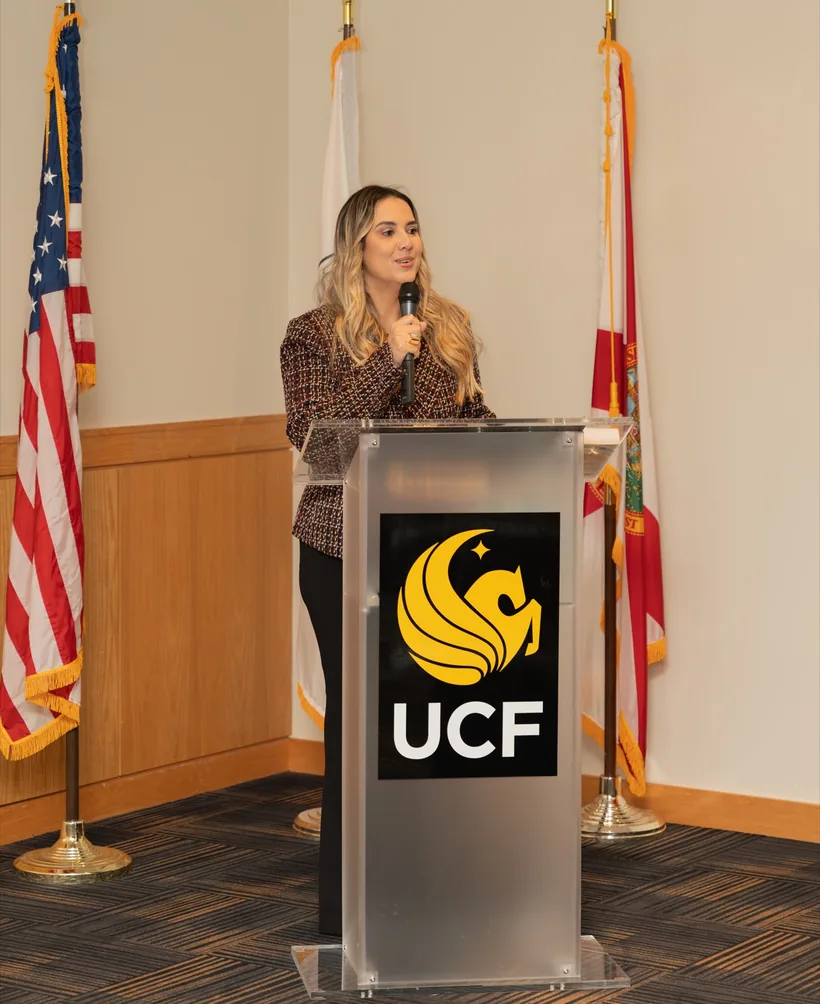 Cristiane Barreto falando em um púlpito com a marca da UCF, na University of Central Florida, entre as bandeiras dos Estados Unidos e da Flórida