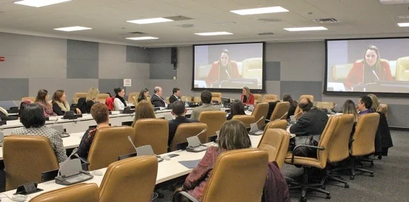 Cristiane Barreto falando a uma plateia sentada em uma sala de conferências da sede das Nações Unidas, com a fala dela projetada em dois telões acima do salão