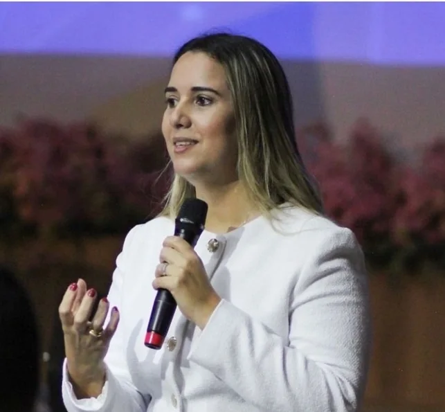 Cristiane Barreto falando com microfone de mão na Western Connecticut State University, com arranjos de flores ao fundo do palco